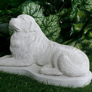 May include: A white, detailed sculpture of a dog in a resting pose. The dog is lying down on a rectangular base, with its head raised and looking upwards. The sculpture is set against a backdrop of green foliage and grass.