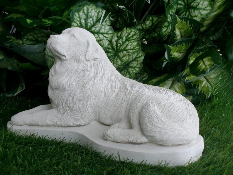 May include: A white, detailed sculpture of a dog in a resting pose. The dog is lying down on a rectangular base, with its head raised and looking upwards. The sculpture is set against a backdrop of green foliage and grass.