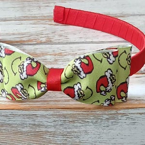 May include: A festive headband featuring a red band and a bow tie. The bow tie has a light green background with a pattern of Santa Claus hats. The headband is a fun accessory for the holiday season.
