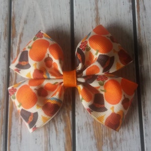 May include: A Thanksgiving-themed hair bow with a white background and orange pumpkins, brown pilgrim hats, and fall leaves. The bow is tied with a bright orange ribbon.