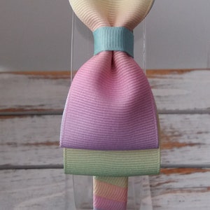 May include: A pastel rainbow headband with a large bow. The bow is made of multiple layers of fabric in shades of blue, green, yellow, pink, and light green.