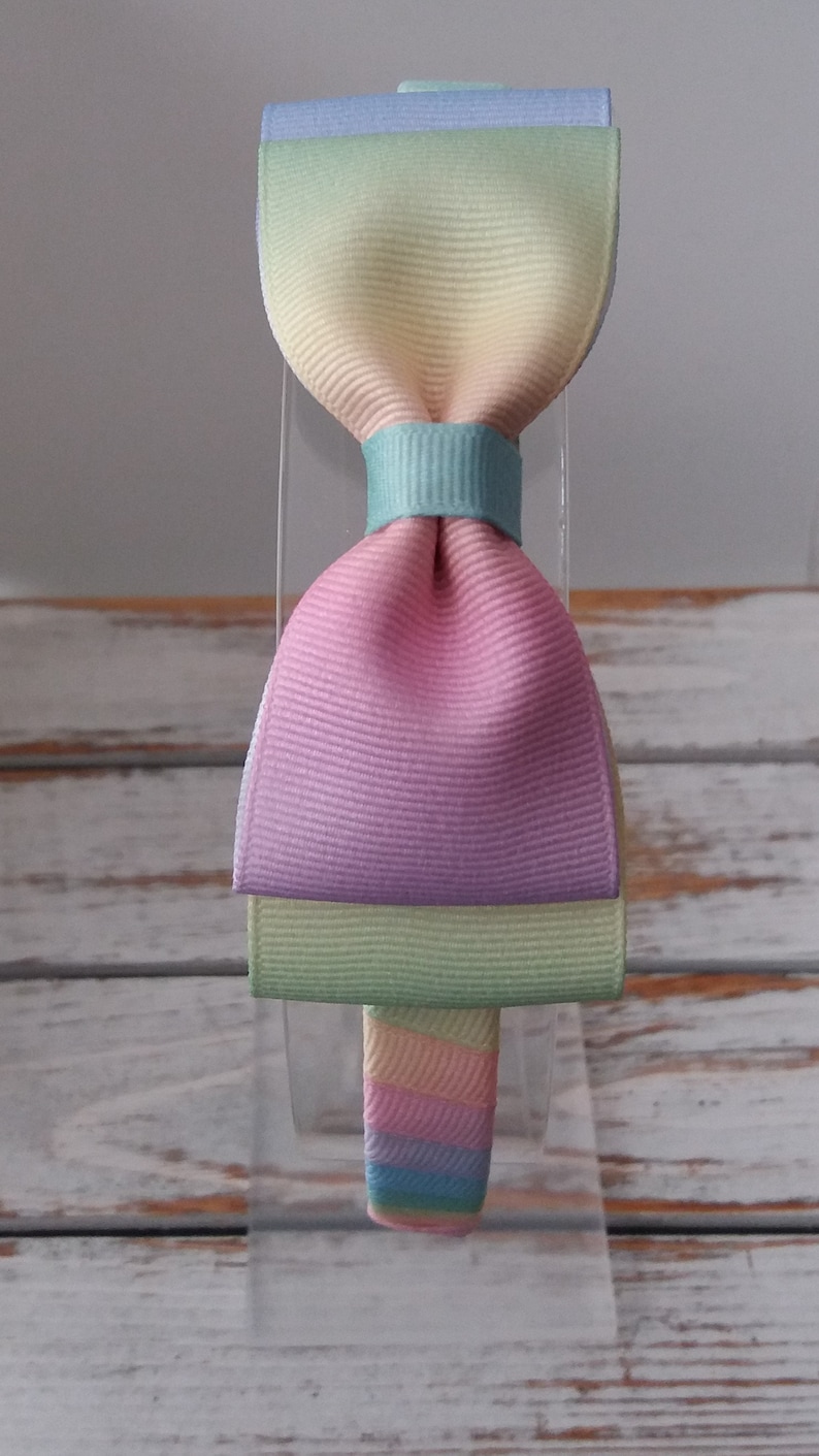 May include: A pastel rainbow headband with a large bow. The bow is made of multiple layers of fabric in shades of blue, green, yellow, pink, and light green.