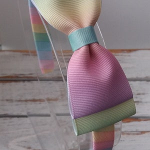 May include: A rainbow headband with a large bow. The bow is made of pastel pink, yellow, green, and blue fabric.
