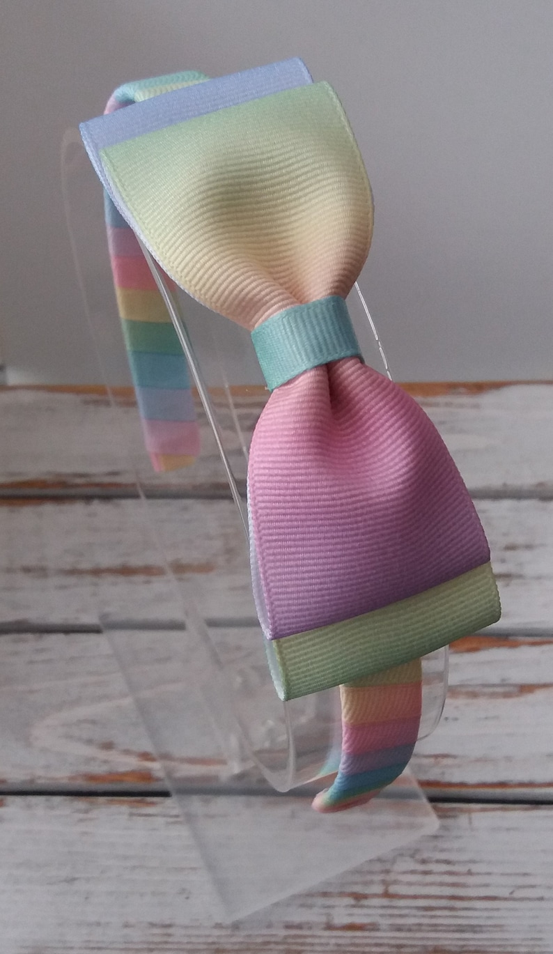 May include: A rainbow headband with a large bow. The bow is made of pastel pink, yellow, green, and blue fabric.