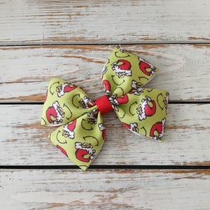 May include: A festive hair bow featuring a green background with a repeating pattern of a smiling face wearing a Santa hat. The bow is tied with a red ribbon in the center.