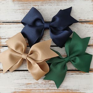May include: Four large, handmade hair bows in navy blue, tan, and forest green. The bows are made of a ribbed grosgrain ribbon and are arranged on a white-washed wooden surface. Each bow is approximately 5 inches wide.