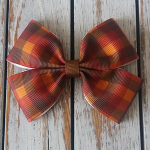 May include: A brown and orange plaid bow with white backing. The bow is tied in a knot and is ready to be attached to a hair clip or headband.