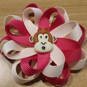 May include: A pink and white ribbon bow with a brown monkey face button in the center. The monkey has a surprised expression.
