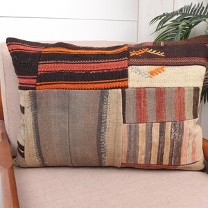 May include: Rectangular patchwork pillow with a variety of colors and patterns. The pillow features sections of brown, orange, beige, and gray fabrics. The pillow is resting on a light pink chair.