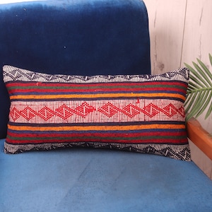 May include: A colourful striped lumbar pillow with a geometric pattern. The pillow is on a blue velvet chair with wooden arms.
