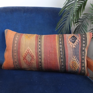 May include: Rectangular pillow with a woven design in shades of orange, red, and brown, featuring diamond and striped patterns. The pillow is placed on a blue chair with wooden armrests, creating a warm contrast.