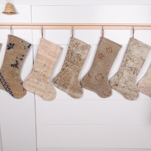 May include: Seven beige and brown Christmas stockings hang from a wooden rod. The stockings have various patterns and textures, including floral and geometric designs. They are suspended against a white wall, with a small bell and greenery in the background.