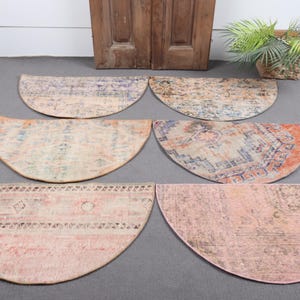 May include: Six half-circle rugs in various colors and patterns are displayed on a gray surface. The rugs feature faded designs in shades of blue, orange, pink, and beige. A wooden door and a potted plant are in the background.