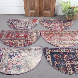 May include: Six half-circle rugs with intricate, faded patterns in shades of red, blue, and cream. The rugs are arranged on a gray surface, with a wooden door and a potted plant in the background. The rugs have a vintage, distressed look.