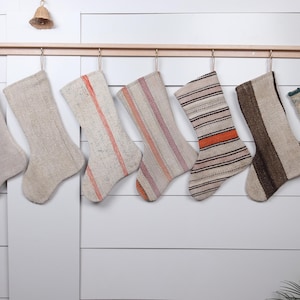 May include: Eight handmade Christmas stockings hanging on a wooden rod. The stockings are made of a natural fabric with various patterns and colors, including beige, brown, orange, and pink.