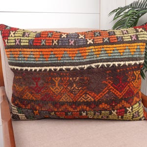 May include: Rectangular decorative pillow with a woven, patterned design. The pillow features a mix of colors including orange, brown, red, and blue. The pillow is resting on a beige chair.