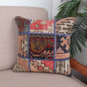 May include: A square decorative pillow with a patchwork design. The pillow features various geometric patterns in shades of red, blue, brown, and beige. The pillow is placed on a beige sofa.