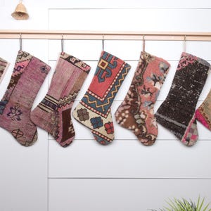 May include: Seven colorful Christmas stockings made from repurposed rugs hang on a wooden rod. The stockings are in shades of pink, brown, beige, and blue with intricate patterns.