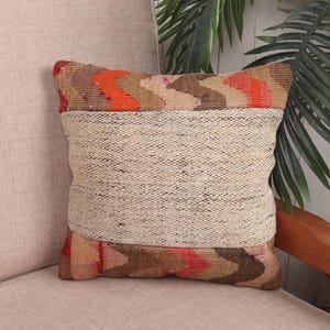 Op de afbeelding: Vierkant decoratief kussen met een geweven beige midden en patronen aan de boven- en onderkant. Het bovenste gedeelte heeft oranje, bruine en beige zigzagpatronen. Het kussen ligt op een beige gestoffeerde stoel.