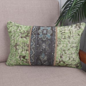 Pode incluir: Almofada decorativa retangular com um design estampado em verde e cinza. A almofada apresenta um painel central com um padrão geométrico em tons de cinza e azul, emoldurado por um fundo verde claro. A almofada está colocada numa cadeira de tom neutro.