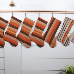 May include: Eight striped Christmas stockings hanging on a wooden rod. The stockings are made of woven fabric and have a mix of orange, brown, and white stripes.