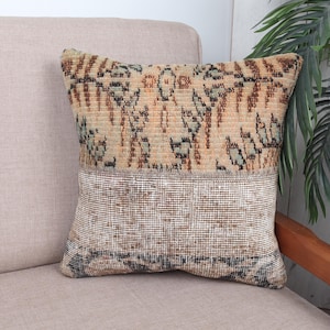 May include: A decorative square pillow with a two-tone design. The top section features a patterned design in shades of brown, green, and beige. The bottom section has a woven texture in beige and brown. The pillow is placed on a light beige sofa.