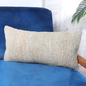 May include: A light gray rectangular pillow with a textured woven fabric sits on a blue velvet armchair. The armchair has wooden arms and a wooden base.