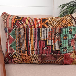 May include: Rectangular decorative pillow with a patchwork design. The pillow features a mix of colors including red, orange, green, and brown. The pillow is placed on a beige chair, with a wooden armrest visible.