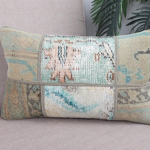 May include: A rectangular patchwork cushion with a vintage design. The cushion displays a mix of muted colours, including beige, teal, and faded pink, arranged in a geometric pattern. It is placed on a neutral-toned sofa, suggesting a cosy home decor item.