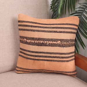 May include: A square, peach-colored pillow with horizontal dark brown stripes. The pillow has a woven texture and is placed on a beige sofa. The pillow is a decorative item.