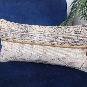 Pode incluir: Almofada retangular decorativa com um design de tapete vintage. A almofada apresenta um fundo bege com padrões florais azuis e castanhos. Uma borda dourada separa as seções superior e inferior. Está colocada numa cadeira de veludo azul.