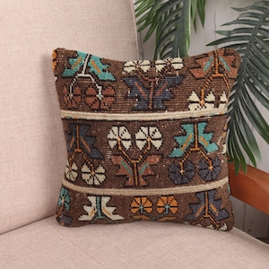 May include: A square, brown decorative pillow with a woven, floral pattern. The pillow features a repeating design of stylized flowers in white, teal, orange, and navy blue. The pillow is placed on a light pink upholstered surface.