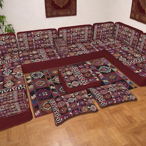 Puede incluir: Un juego de sofá seccional burdeos con un diseño estampado en tonos morados y crema. El conjunto incluye un sofá esquinero, una otomana rectangular y varios cojines. El sofá y la otomana están colocados sobre una alfombra a juego.