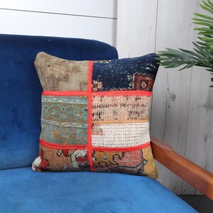 May include: Square patchwork pillow with a variety of patterned fabric pieces in beige, blue, and red. The pillow is on a blue chair, with a green plant in the background. The pillow is approximately 16 inches square.