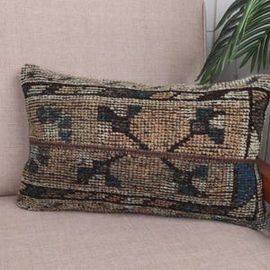 Puede incluir: Cojín decorativo rectangular con un diseño tejido y texturizado. El cojín presenta un patrón en tonos marrones, beige y azules. Se coloca sobre una silla rosa claro. El cojín mide aproximadamente 30 cm de largo y 20 cm de ancho.