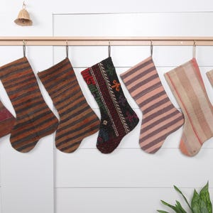 May include: Seven colorful striped stockings hang on a wooden rod. The stockings are made of woven fabric and have a variety of patterns and colors, including red, brown, pink, and beige.