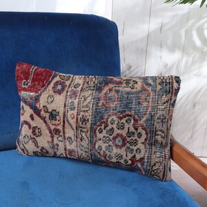 May include: Rectangular decorative pillow with a vintage rug design. The pillow features a beige background with intricate patterns in red, blue, and beige. It rests on a blue velvet armchair, with a plant in the background.