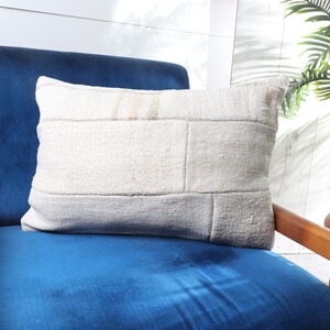May include: A white, off-white, and beige patchwork pillow sits on a blue velvet armchair with wooden arms. The chair is in front of a white wood paneled wall.
