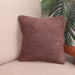 May include: A square, reddish-brown decorative cushion with a textured weave. The cushion has a stitched detail along the top edge. It is placed on a beige sofa, with green plants in the background. The cushion is approximately 40 cm square.