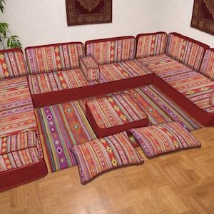 Puede incluir: Un sofá modular en esquina con una tela estampada vibrante en tonos rojos, naranjas, morados y amarillos. El sofá tiene un perfil bajo y un marco rojo oscuro. Incluye cojines a juego y una alfombra con un diseño similar.