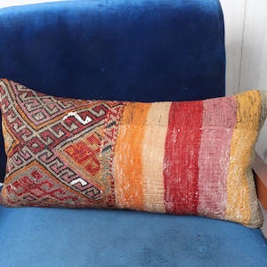 Könnte beinhalten: Ein langes, rechteckiges Kissen mit einem farbenfrohen, gemusterten Design. Das Kissen ist aus gewebtem Stoff mit einem Mix aus roten, orangen, gelben und weißen Streifen und geometrischen Mustern. Das Kissen liegt auf einem blauen Samtstuhl.