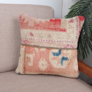 May include: A decorative pillow with a vintage rug design. The pillow features a cream-colored background with red and blue geometric patterns. It is placed on a beige sofa, with a plant in the background.