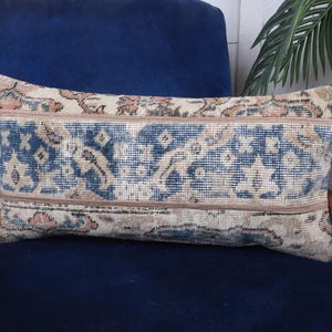 May include: Rectangular decorative pillow with a vintage rug design. The pillow features a central blue field with a repeating pattern, framed by beige and brown borders. The pillow is placed on a dark blue velvet chair.