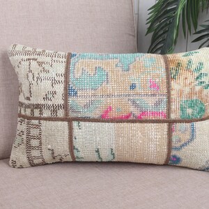May include: Rectangular patchwork pillow with a beige background and various colorful geometric patterns. The pillow features a mix of beige, blue, pink, and green hues, with brown stitching. The pillow is placed on a light pink surface.