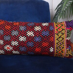 May include: Rectangular decorative pillow with a geometric pattern in red, white, purple, and orange. The pillow is placed on a blue chair. The pillow's design features diamond shapes and floral motifs, adding a touch of cultural flair.