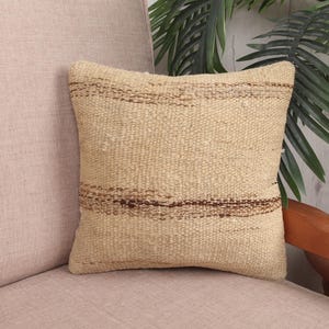 May include: A square, beige woven pillow with horizontal brown stripes. The pillow is placed on a light pink upholstered chair. The pillow has a textured appearance and a neutral color palette, suitable for various home decor styles.