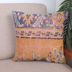 May include: A decorative square pillow with a textured, vintage-style design. The pillow features a color palette of orange, blue, and brown. The pillow is placed on a beige sofa, with a wooden chair and green plant in the background.