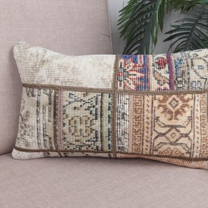 May include: Rectangular decorative pillow with a patchwork design. The pillow features a cream-colored top and a patchwork of patterned fabric squares in shades of beige, brown, blue, and red. The pillow is placed on a light pink sofa.