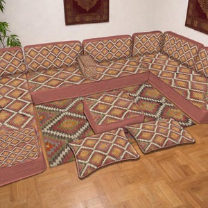 May include: A modular sofa set with a geometric diamond pattern in shades of orange, yellow, and white on a red-brown background. The set includes multiple seat cushions, backrests, and throw pillows, arranged on a wooden floor.