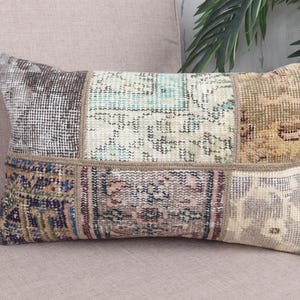 May include: Rectangular patchwork pillow with a variety of muted colors, including beige, brown, and blue. The pillow has a vintage, distressed look, with each patch featuring a different pattern. The pillow is placed on a light pink upholstered surface.
